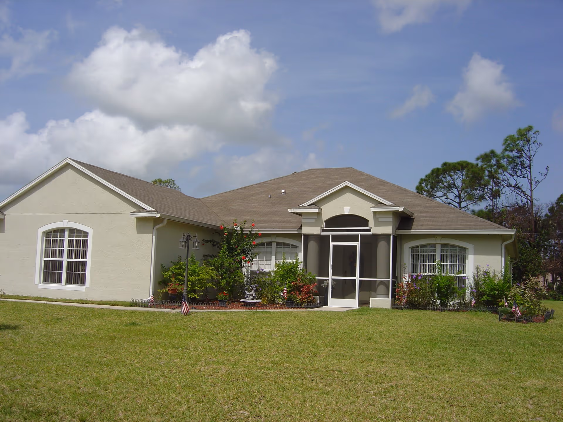 Single-story light-colored house with a screened front porch, lawn and landscaping under a blue sky.