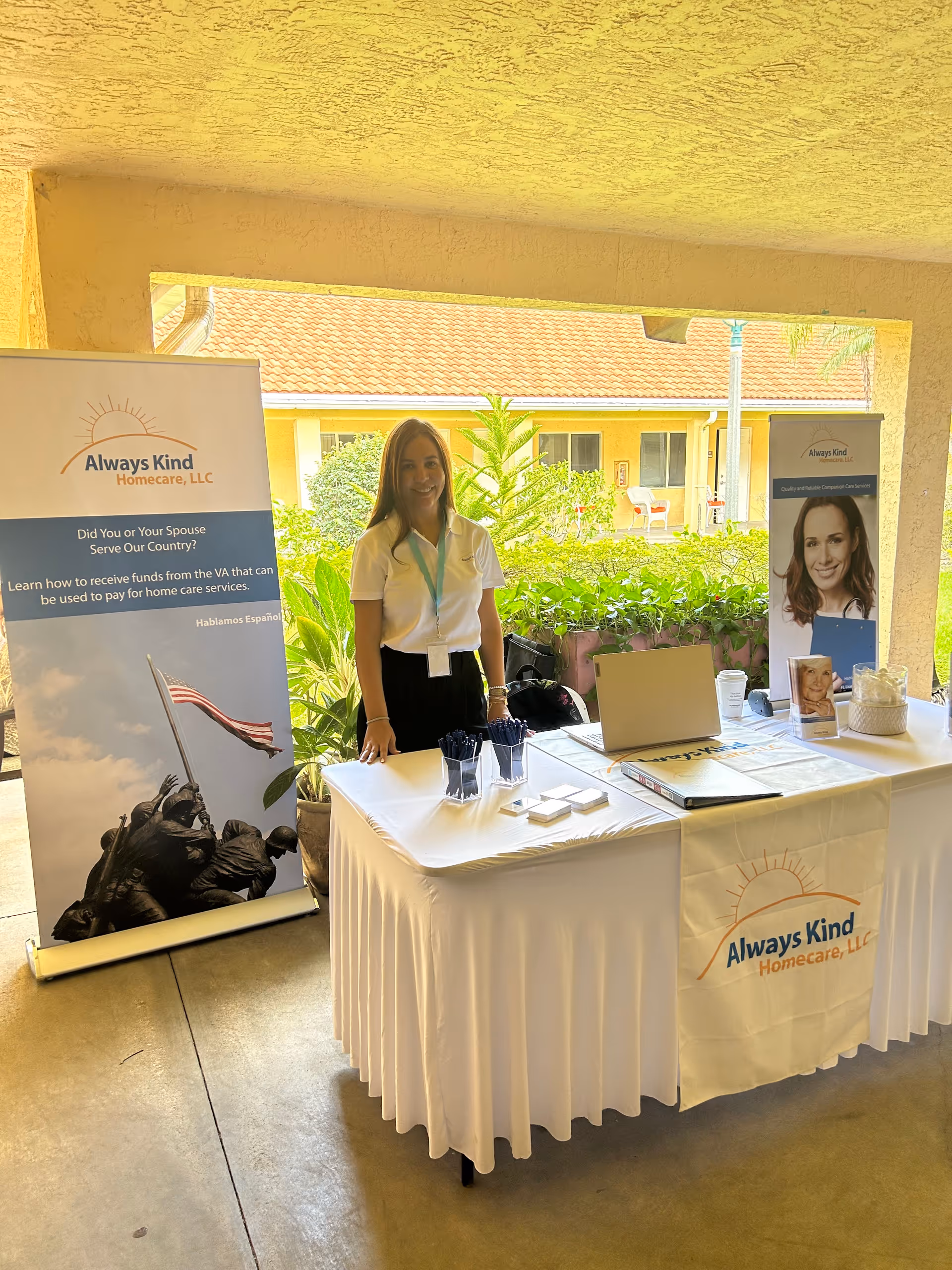 A staff member stands behind an Always Kind HomeCare booth with banners and a laptop.