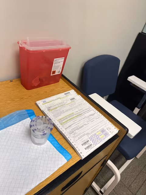 Desk with medical forms, red sharps container, and blue chair in a clinic interior.