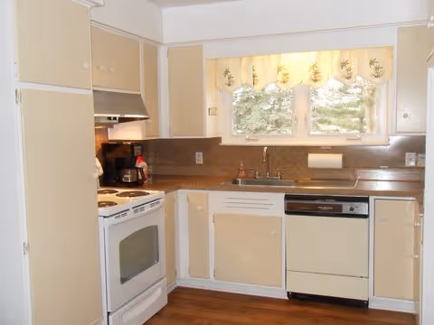 A compact beige kitchen with stove, dishwasher, sink, and a window.