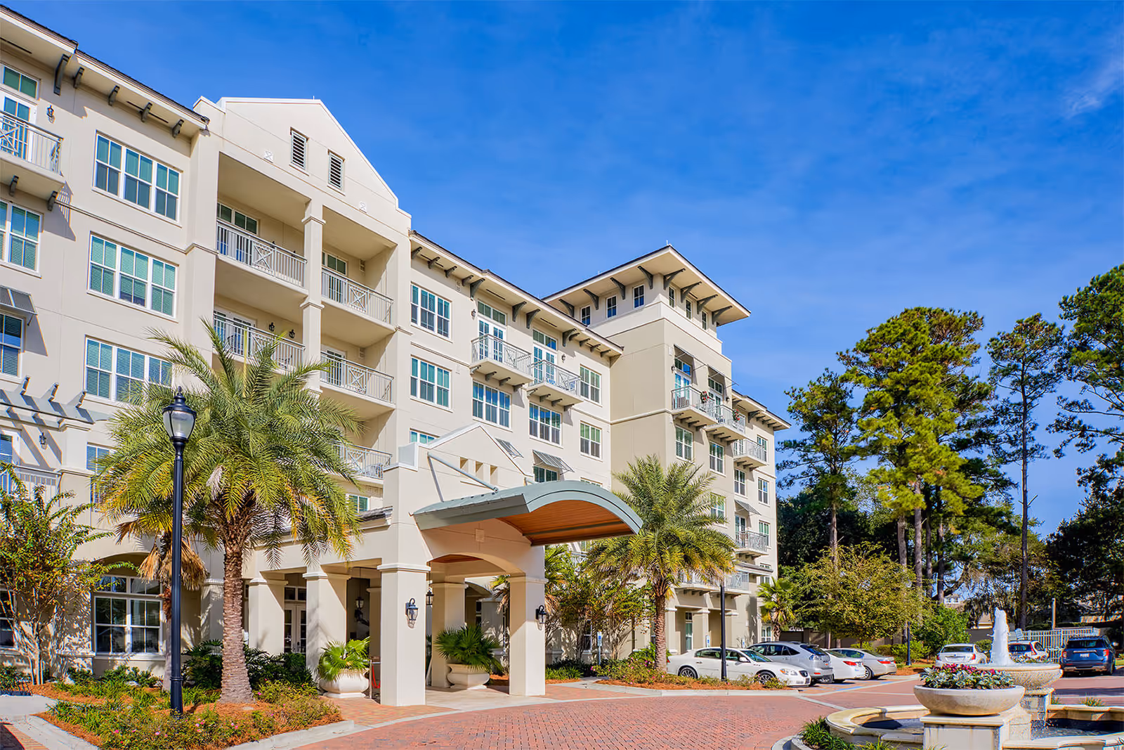 Exterior view of a multi-story senior living facility with balconies, palm trees, a covered entrance, parked cars, and a fountain in the foreground under a clear blue sky.