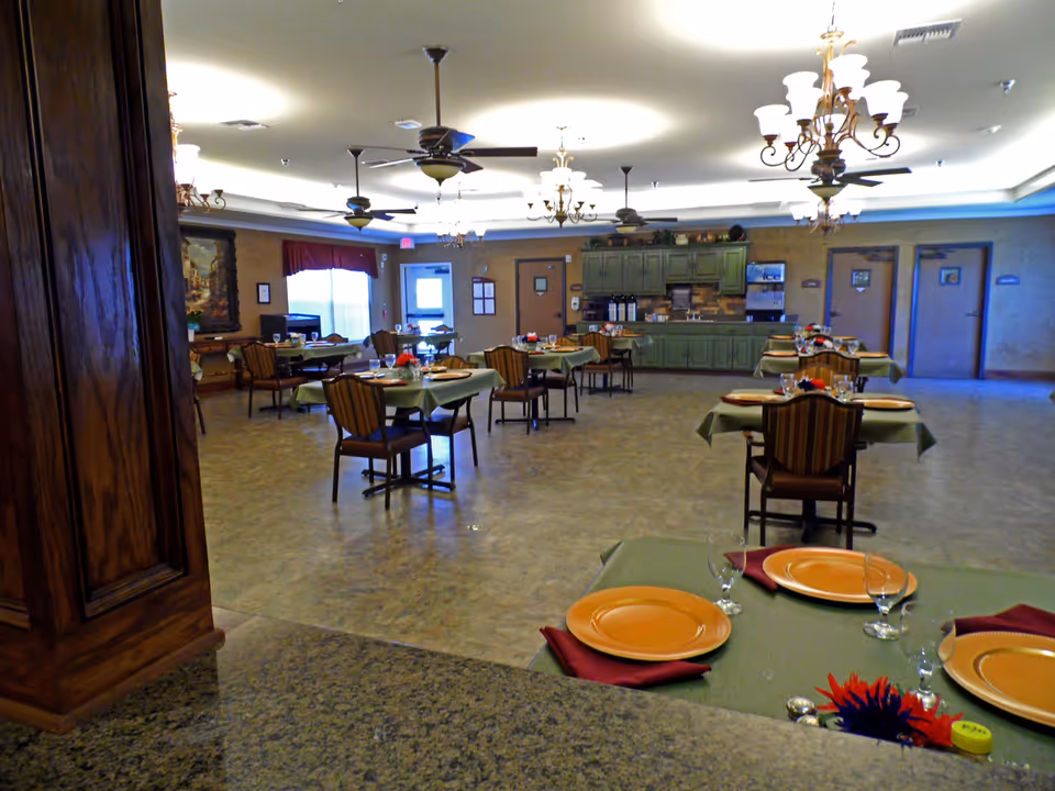 A spacious dining room with multiple tables set with orange plates, glasses, and red napkins. The room features ceiling fans, chandeliers, and green cabinets along the back wall with a beverage dispenser. There are several doors and a window with a red valance in the background.