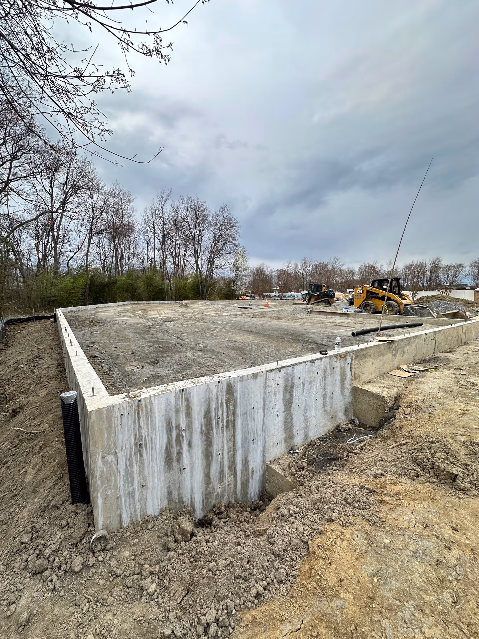 Under-construction concrete foundation with dirt, equipment, and bare trees under a cloudy sky