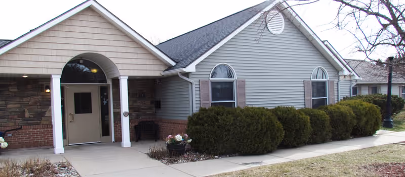 Exterior front view of a single-story assisted living facility building with gray siding, a gabled roof, and an arched entrance supported by white columns. There are shrubs and a small garden area along the sidewalk leading to the entrance.
