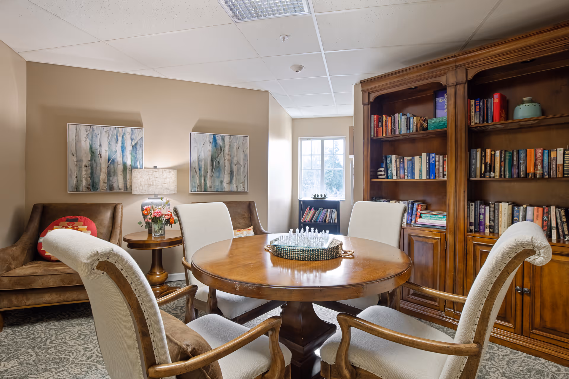 Cozy common room with a round wooden table surrounded by upholstered chairs, armchairs, and a large bookshelf.
