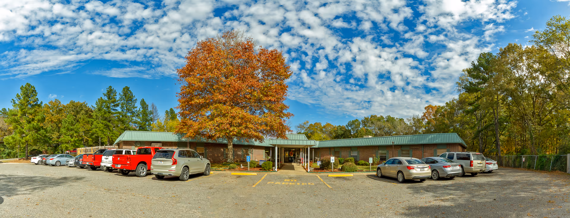 Exterior view of a single-story brick building with a green metal roof, surrounded by trees with autumn foliage. Several cars are parked in the gravel parking lot in front of the building under a partly cloudy blue sky.