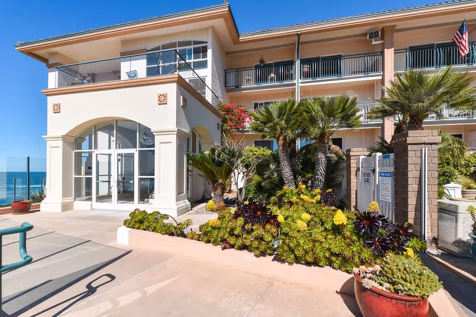 Exterior view of a multi-story senior living facility building with balconies, palm trees, and various plants in front. The building has a light-colored facade with large glass doors and windows. An American flag is visible on the right side, and the ocean can be seen in the background under a clear blue sky.