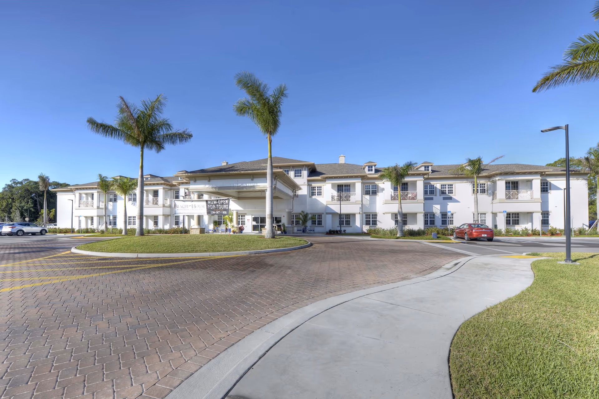 Front exterior view of a two-story senior living facility named Beach House with a circular driveway, palm trees, and a clear blue sky.