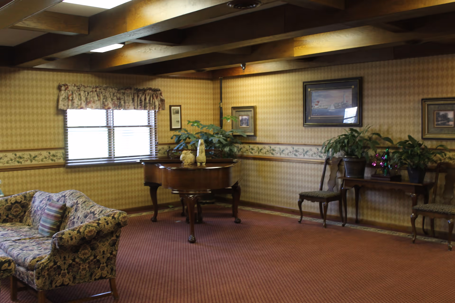 A carpeted common lounge with a baby grand piano, patterned sofa, potted plants, framed artwork, and exposed wooden ceiling beams.