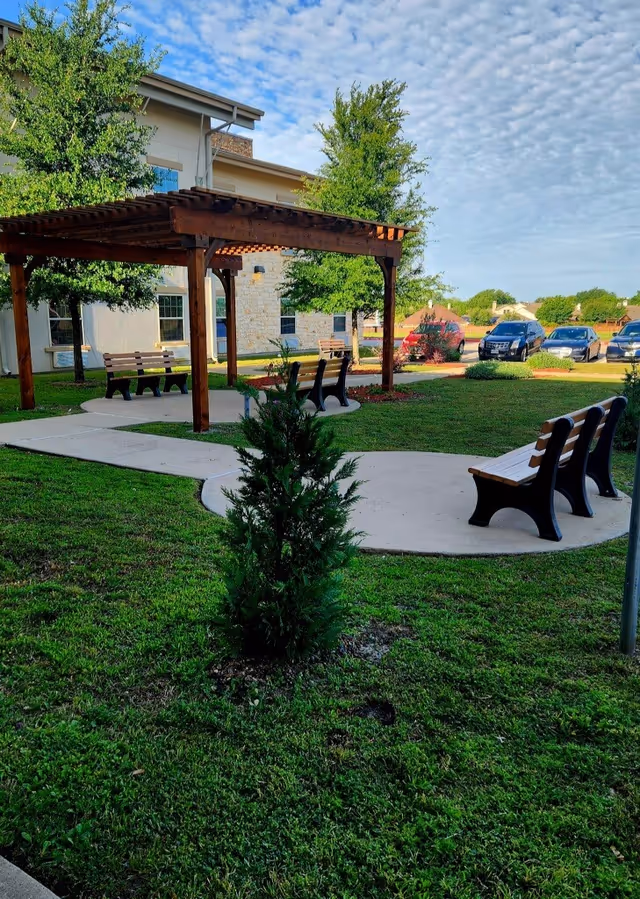 Outdoor courtyard with a wooden pergola, benches along curved paved paths, grassy lawn and a building with parked cars in the background.