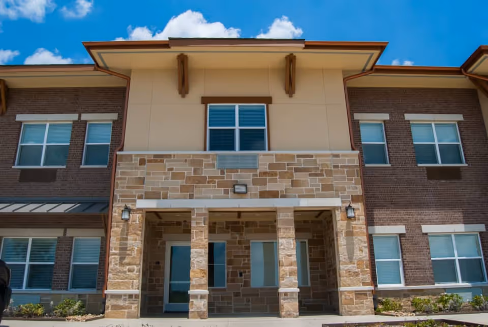 Front exterior view of Cedar Bluff Assisted Living & Memory Care building with a stone and brick facade, multiple windows, and a covered entrance under a clear blue sky with some clouds.