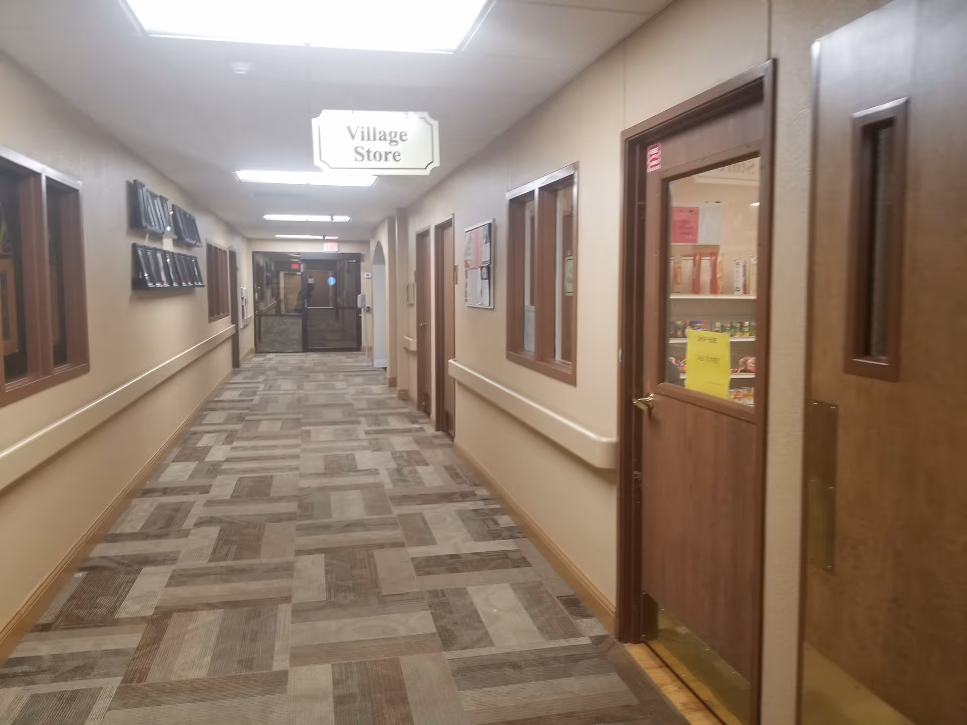 Long beige hallway of a senior living facility with doors, windows, and a Village Store sign.