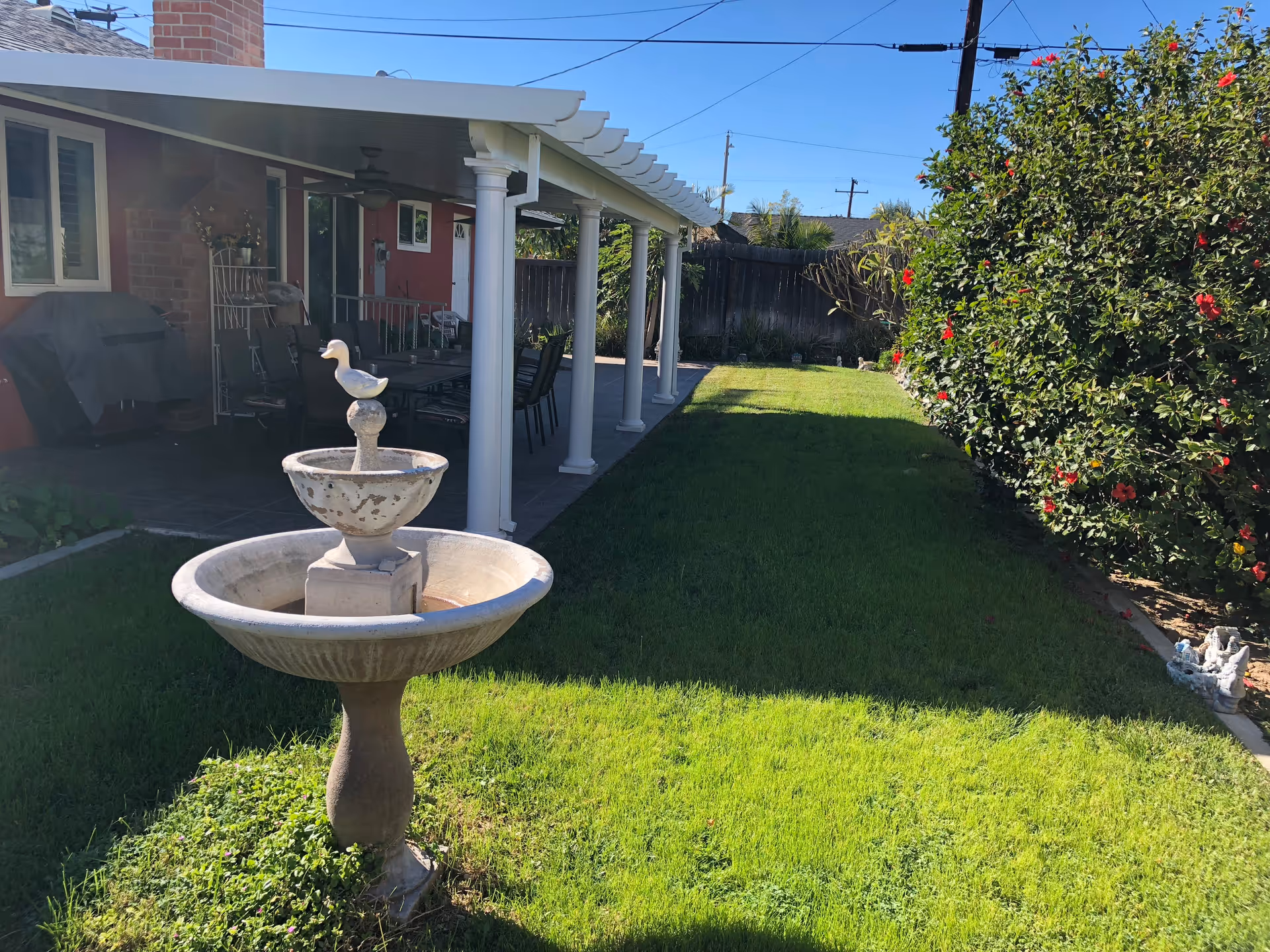 A sunny backyard with a stone fountain, lawn, and a covered patio with chairs.