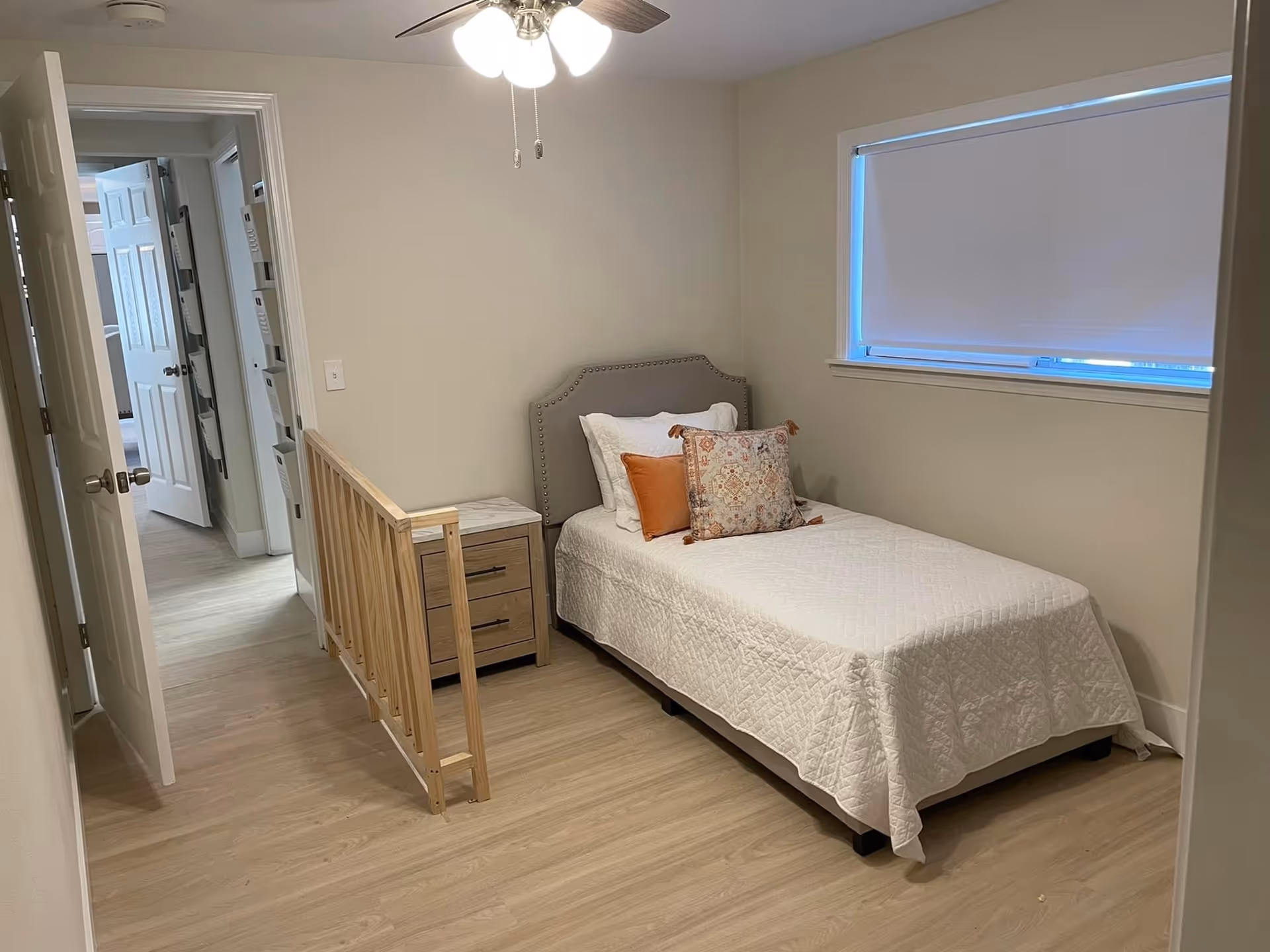 A small bedroom with a single bed, nightstand, window blind, and ceiling fan.