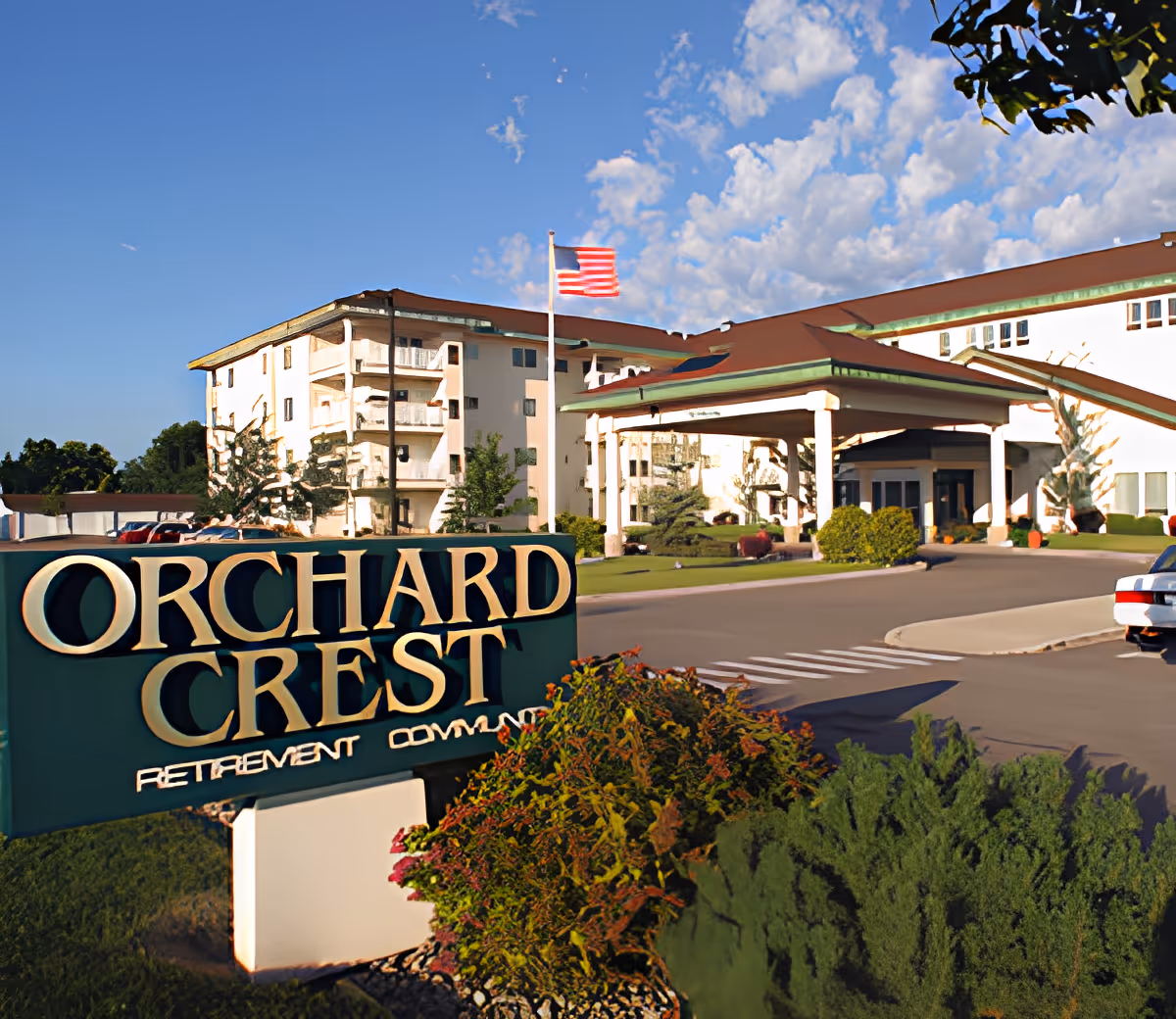 Front entrance and sign for Orchard Crest Retirement Community with a multi-story building, landscaping, and an American flag.