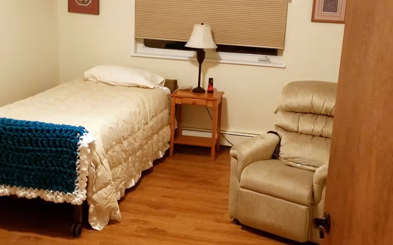 A small bedroom with a single bed covered in a beige quilt and a blue crocheted blanket at the foot. Next to the bed is a wooden nightstand with a lamp and a small red object. A beige recliner chair is positioned near the door. The room has wooden flooring, a window with closed blinds, and light-colored walls with two framed pictures.