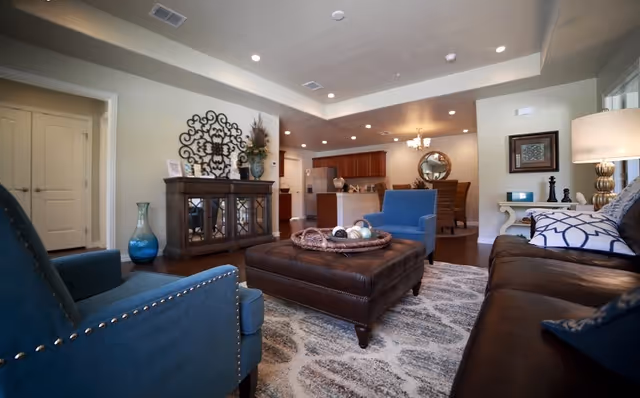 A cozy living room with blue upholstered armchairs, a brown leather sofa, and a large brown leather ottoman in the center. The room features a patterned area rug, a wooden cabinet with decorative items, and a lamp on a side table. In the background, there is a dining area with a round mirror on the wall and a kitchen with wooden cabinets and stainless steel appliances.
