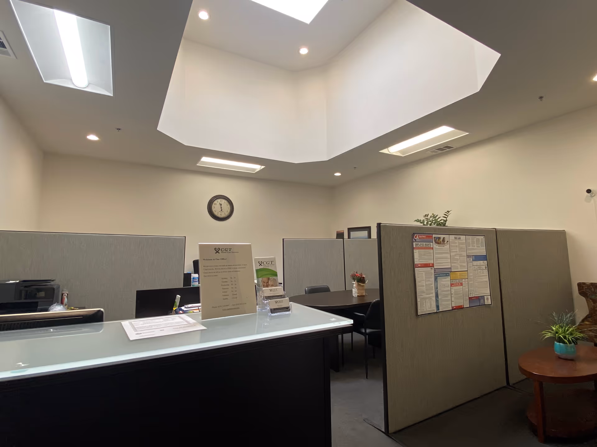 Reception area with cubicles, a front desk, clock, and skylights in a neutral office.
