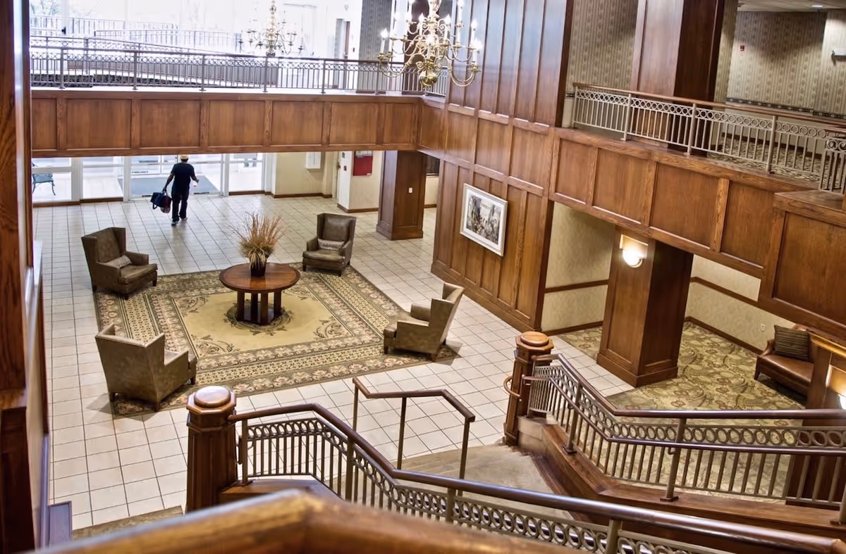 Spacious two-story wood-paneled lobby with seating around a central rug, staircase and railing overlooking the tiled entry.