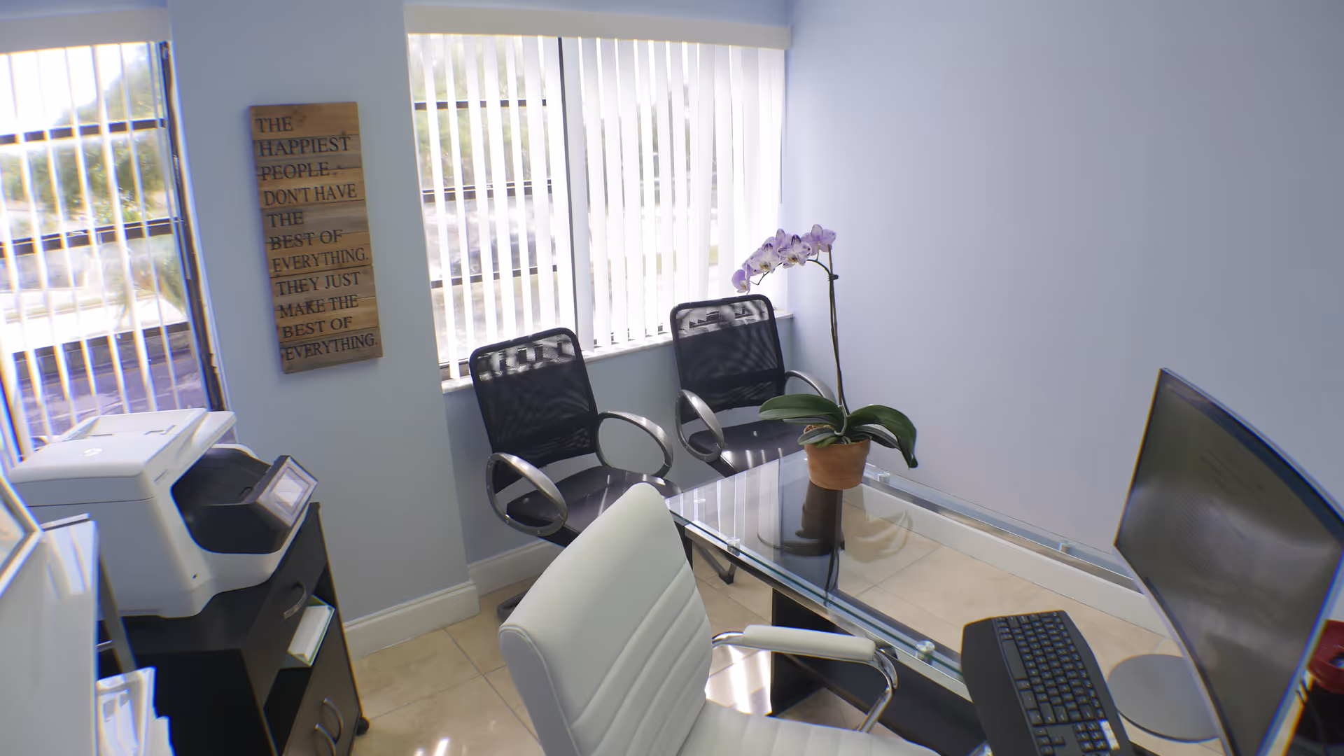 Modern office reception area with glass desk, computer, printer, chairs, and an orchid plant.