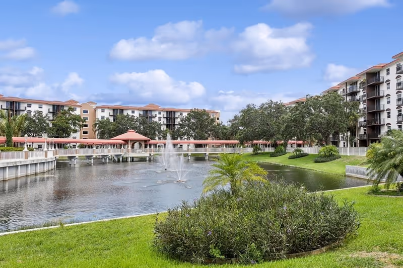 Pond with a central fountain, landscaped lawn and palm shrubs in front of multi-story senior living buildings connected by a covered walkway.