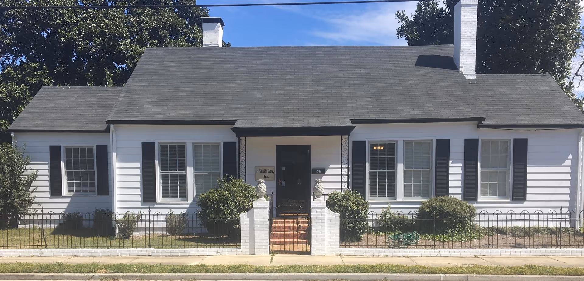 White single-story senior care facility front entrance with black shutters and a fenced yard.