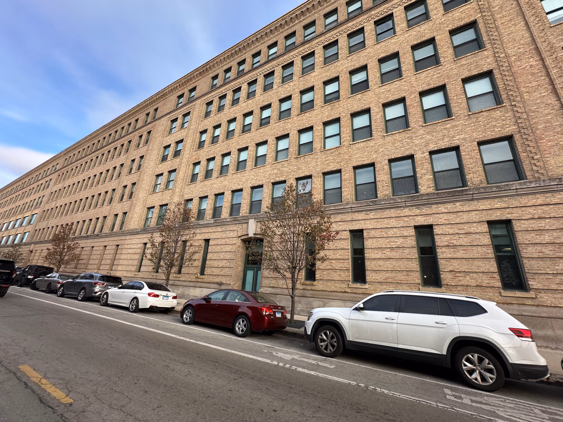Exterior view of a large multi-story brick building with many windows, several parked cars along the street, and a few small trees in front of the building under a partly cloudy sky.