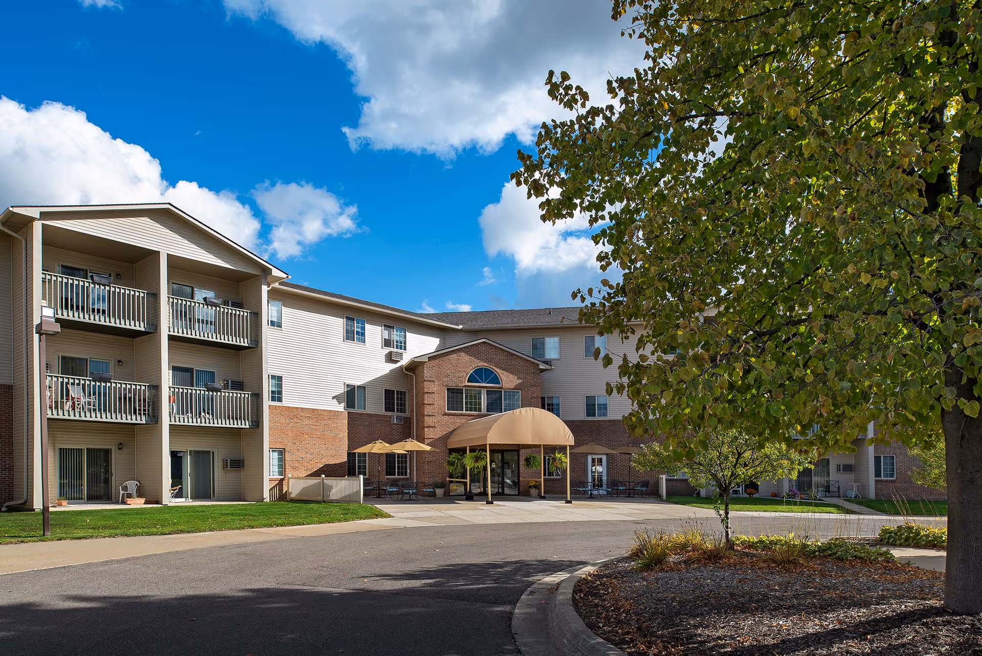 Front exterior of a multi-story senior living building with balconies, a covered main entrance, and a tree-lined driveway under a blue sky.