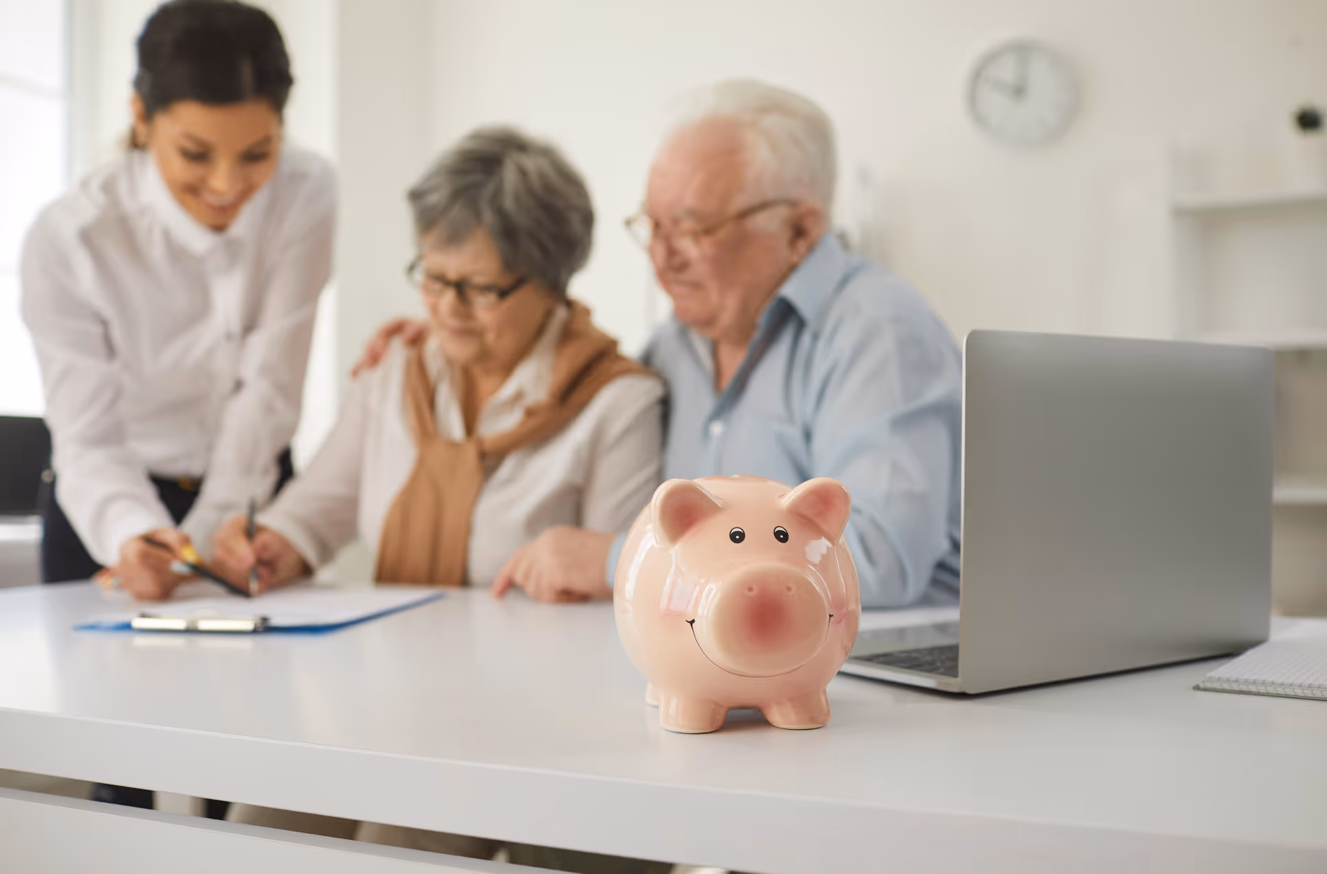 Pink piggy bank sits on a white desk while an elderly couple and caregiver look on.