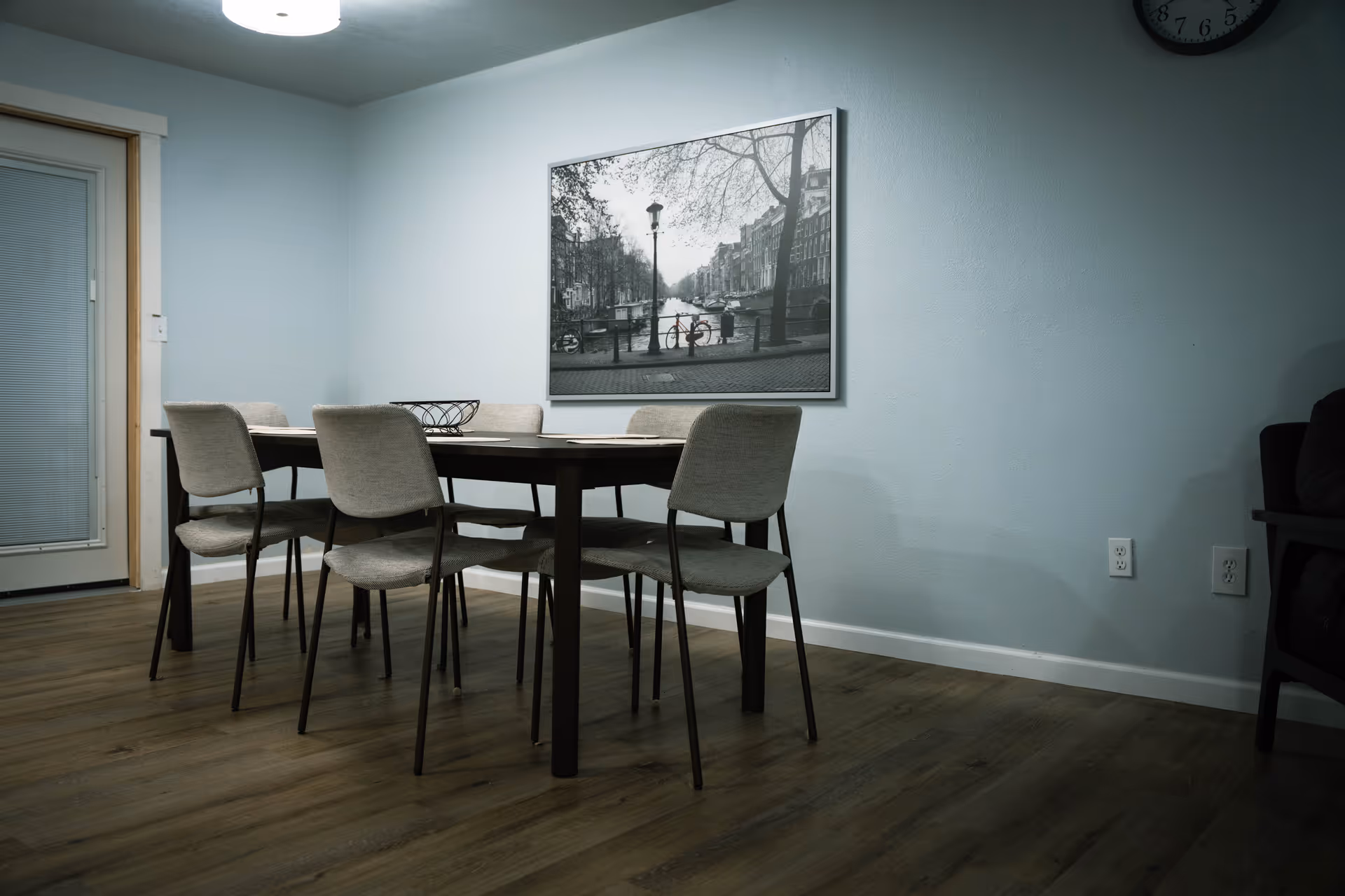 A dining area with a rectangular dark wooden table surrounded by six light gray upholstered chairs. On the wall above the table is a large black and white framed photograph of a canal scene with a bicycle and trees. The room has light blue walls, wood flooring, a clock on the upper right wall, and a door with a window covered by blinds on the left side.