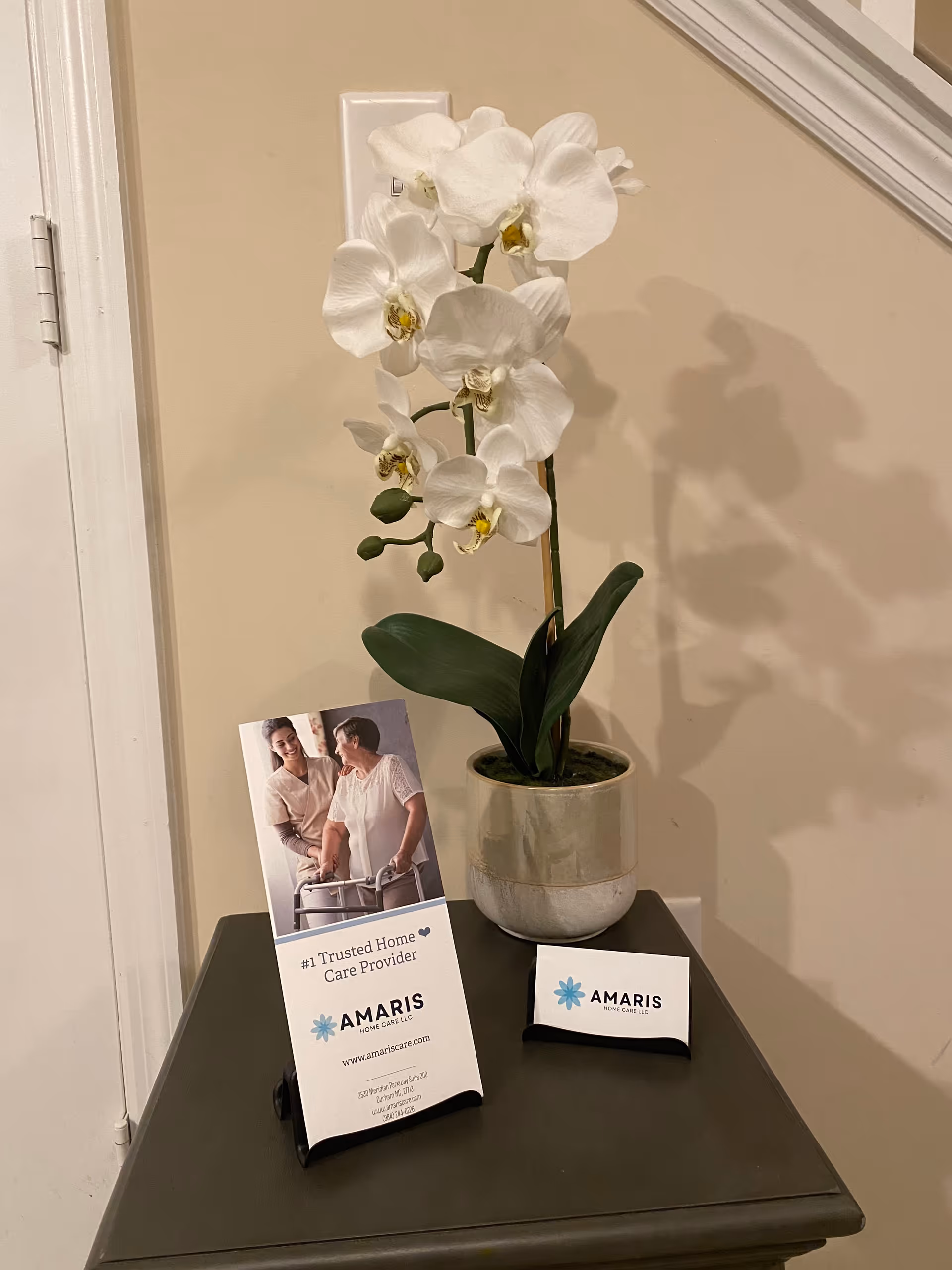 White orchid plant in pot on a dark table beside Amaris Home Care marketing cards