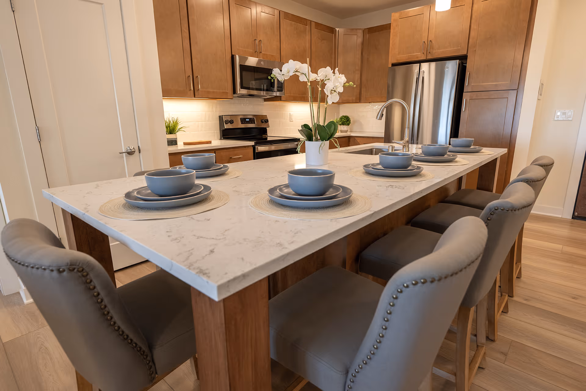 Modern kitchen with a large marble island countertop set with six place settings of gray bowls and plates on round placemats. The island is surrounded by six gray upholstered chairs with nailhead trim. The kitchen features wooden cabinets, a stainless steel refrigerator, oven, and microwave, with a white subway tile backsplash and a potted orchid centerpiece on the island.