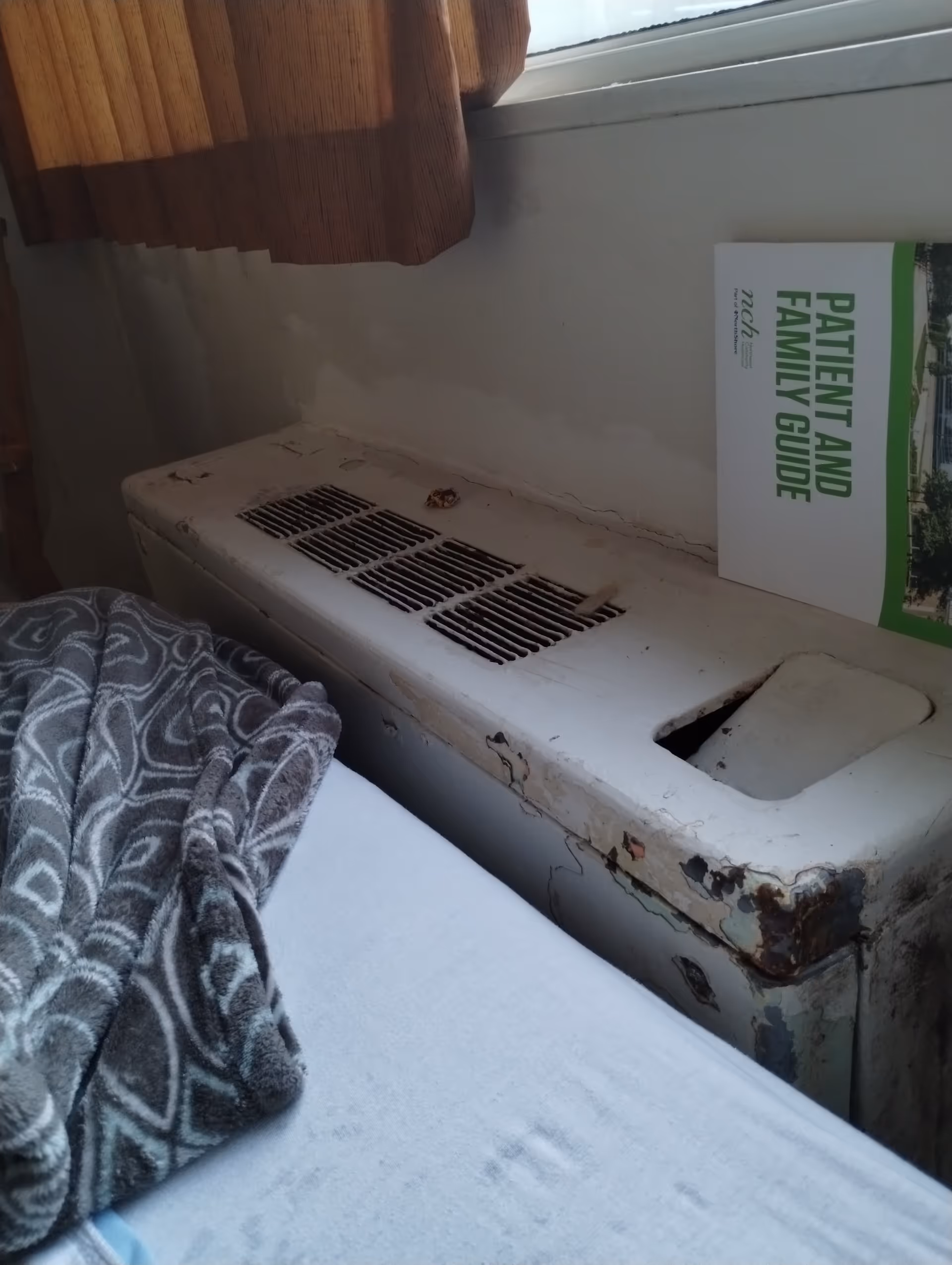 Rusty, worn heater/air conditioner sits beside a bed under a window.
