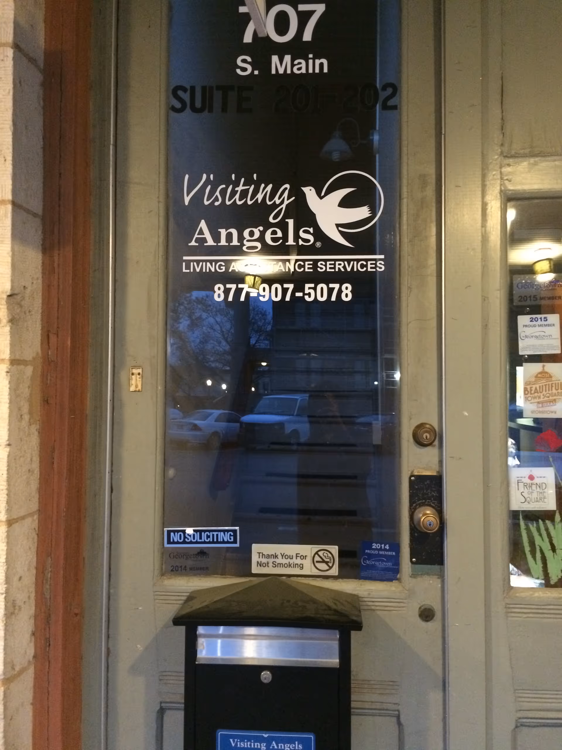 Front entrance door with Visiting Angels signage and phone number, reflecting street scene.