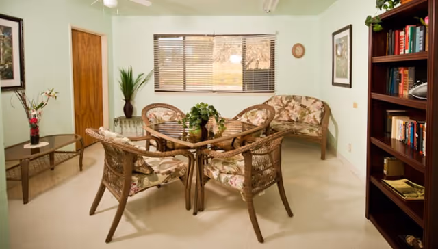 A cozy sitting area in a healthcare facility with a glass-top square table surrounded by four wicker chairs with floral cushions. There is a matching wicker loveseat against the wall, a wooden bookshelf filled with books on the right, a small glass-top side table with a flower vase on the left, and a window with blinds letting in natural light.