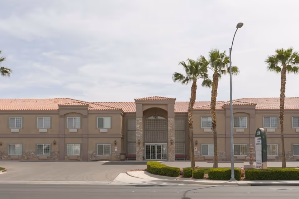 Exterior front view of a two-story senior living facility building with a tiled roof, multiple windows, palm trees, and a sign near the entrance. The sky is partly cloudy.