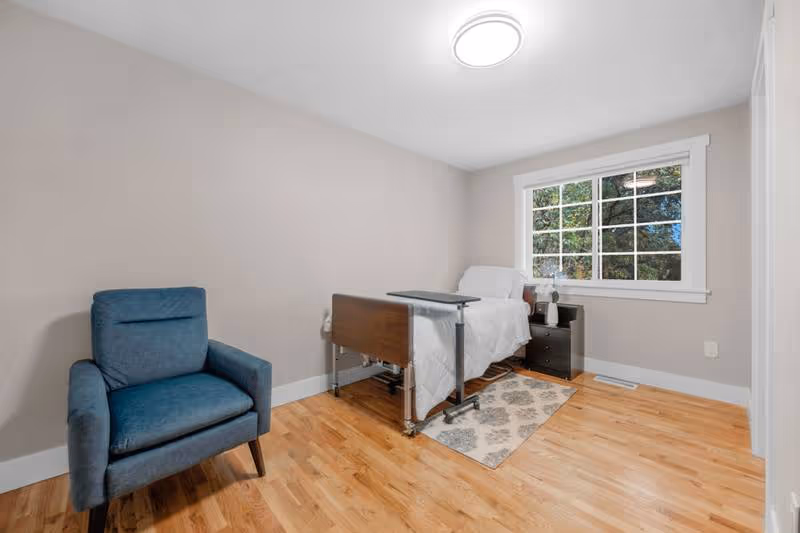 A small, neat bedroom with a bed, blue chair, nightstand, and window.