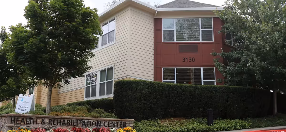 Two-story beige and red senior living building with manicured hedge and Health & Rehabilitation Center sign.