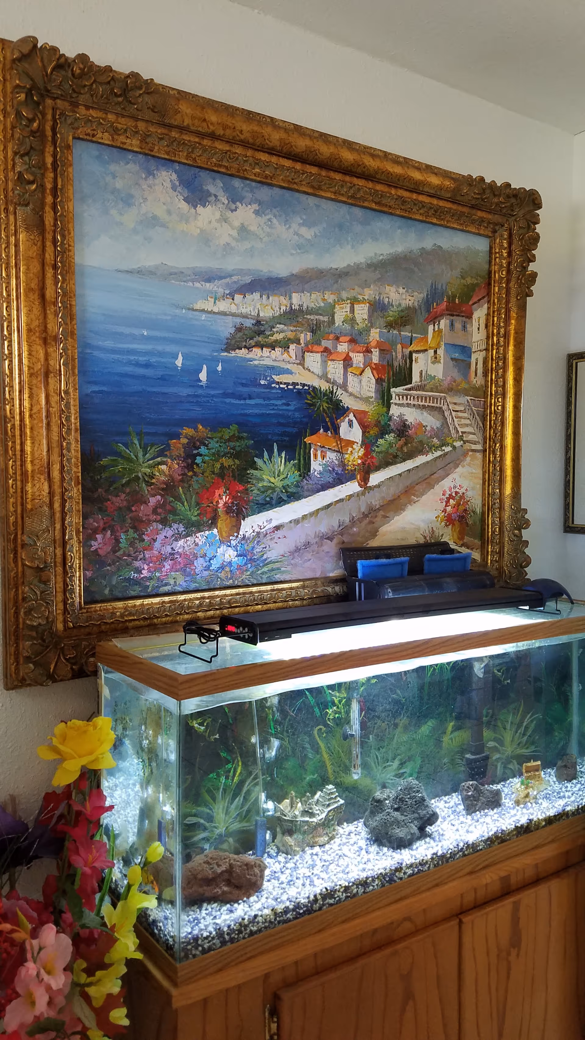 Ornate framed coastal painting above a glass aquarium on a wooden cabinet in a hallway.
