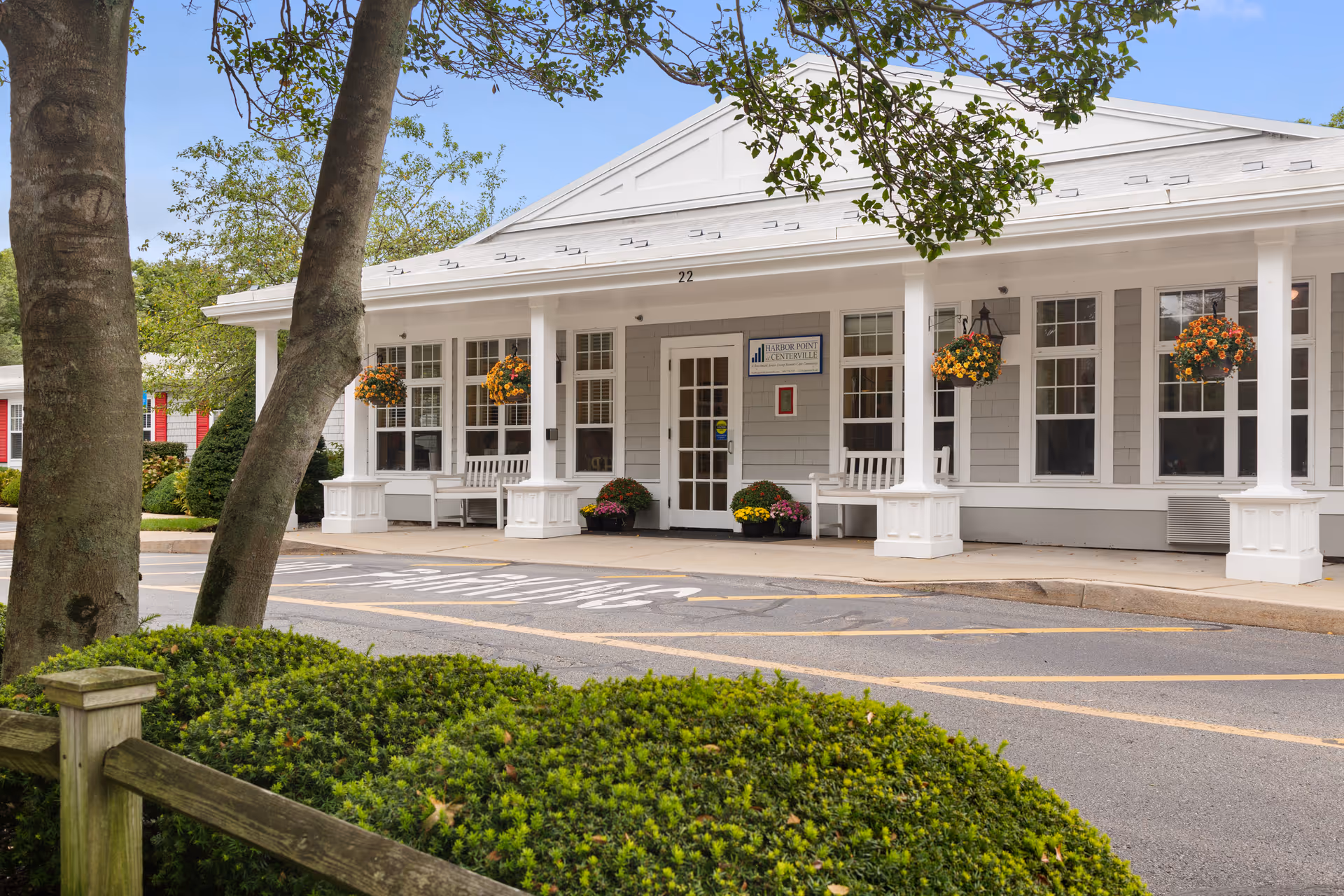 Exterior view of Harbor Point at Centerville building entrance with white columns, hanging flower baskets, benches, and a small garden with trees and bushes in the foreground.
