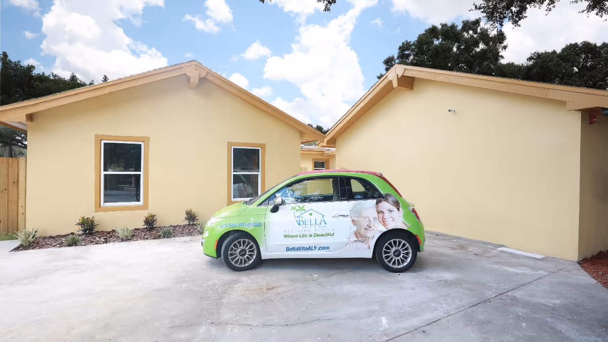 A small green-and-white branded compact car parked on a concrete driveway in front of a yellow single-story building with two windows.