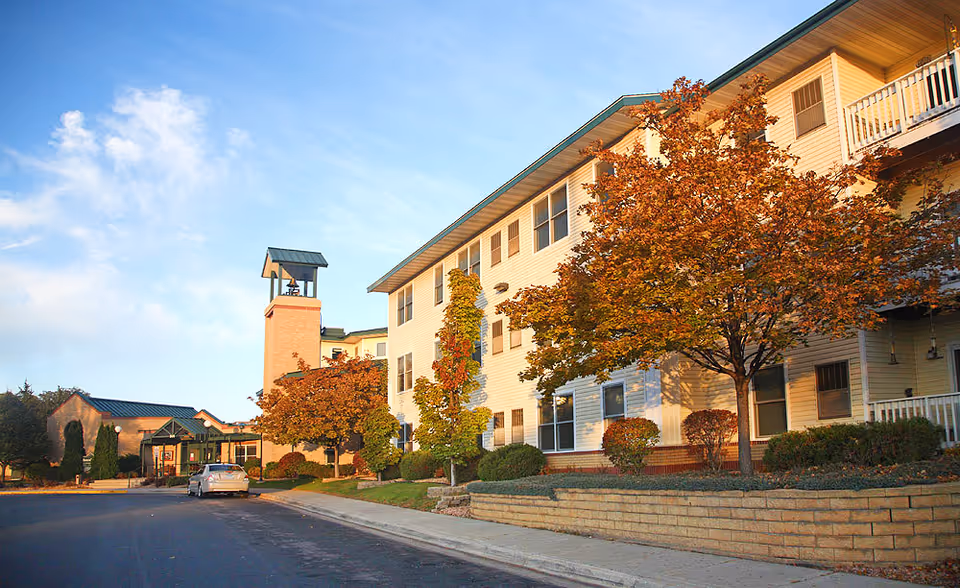 Exterior view of Auburn Homes and Services Waconia building with autumn trees and a clear blue sky. The building is light-colored with multiple windows and a bell tower near the entrance. A car is parked on the driveway in front of the building.
