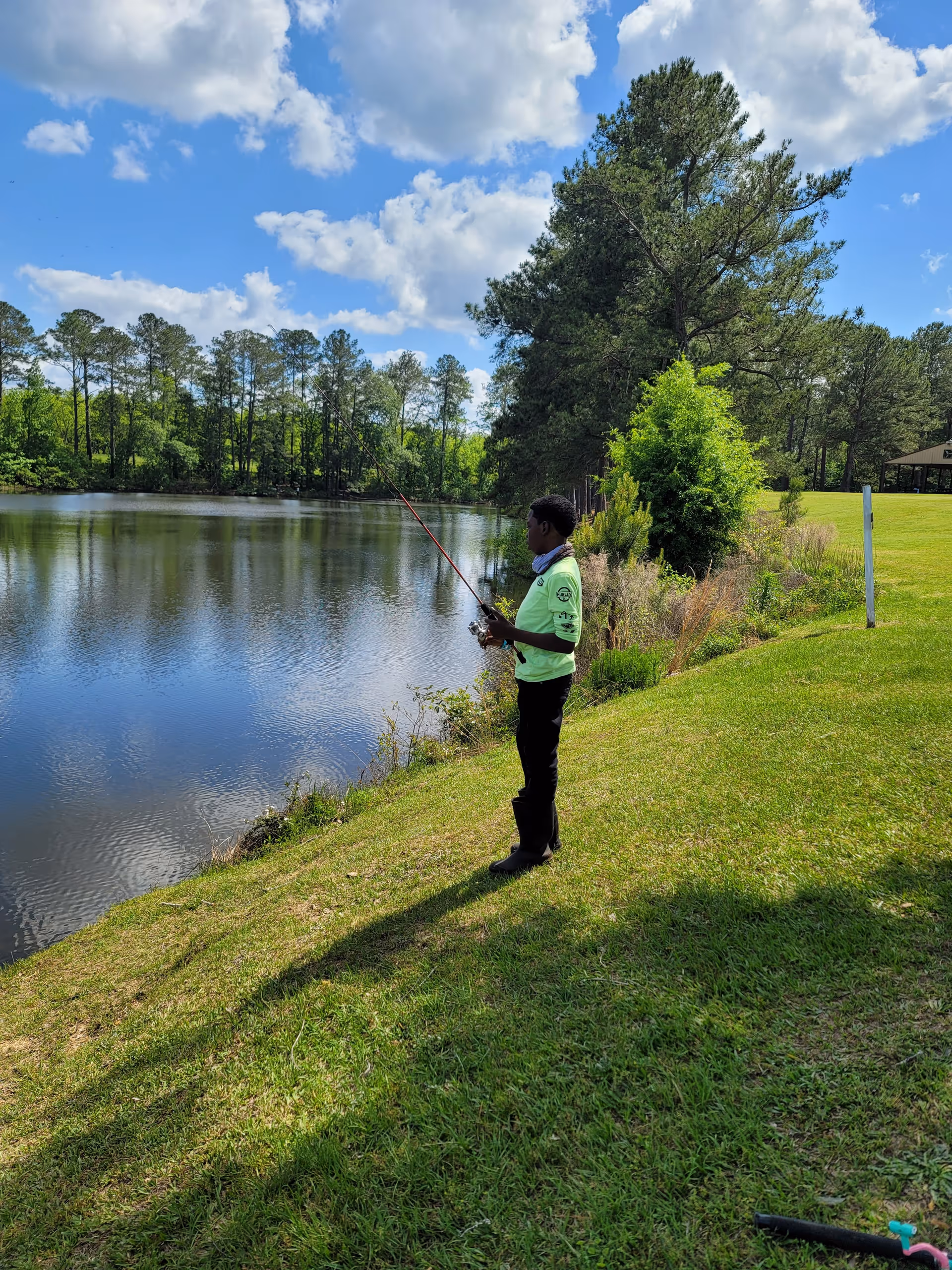 Caregiver in a light uniform fishes by a pond beside trees.