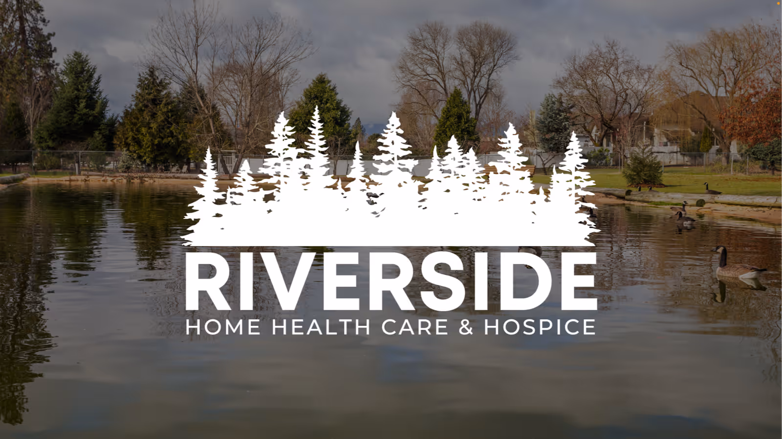 Outdoor lakeside park with trees and a bench, overlaid by Riverside branding.