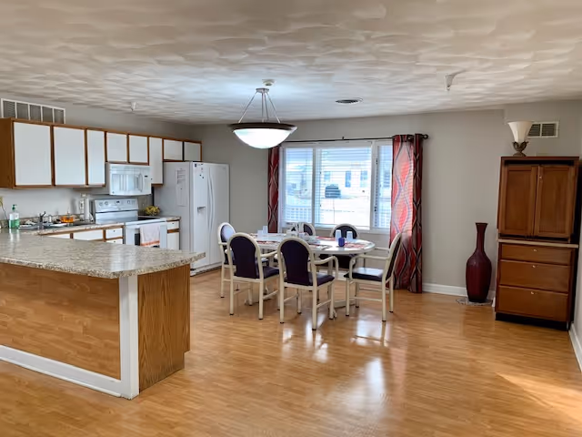 Open communal dining area with a round table and chairs beside a kitchen island, cabinets, and a large window with curtains.