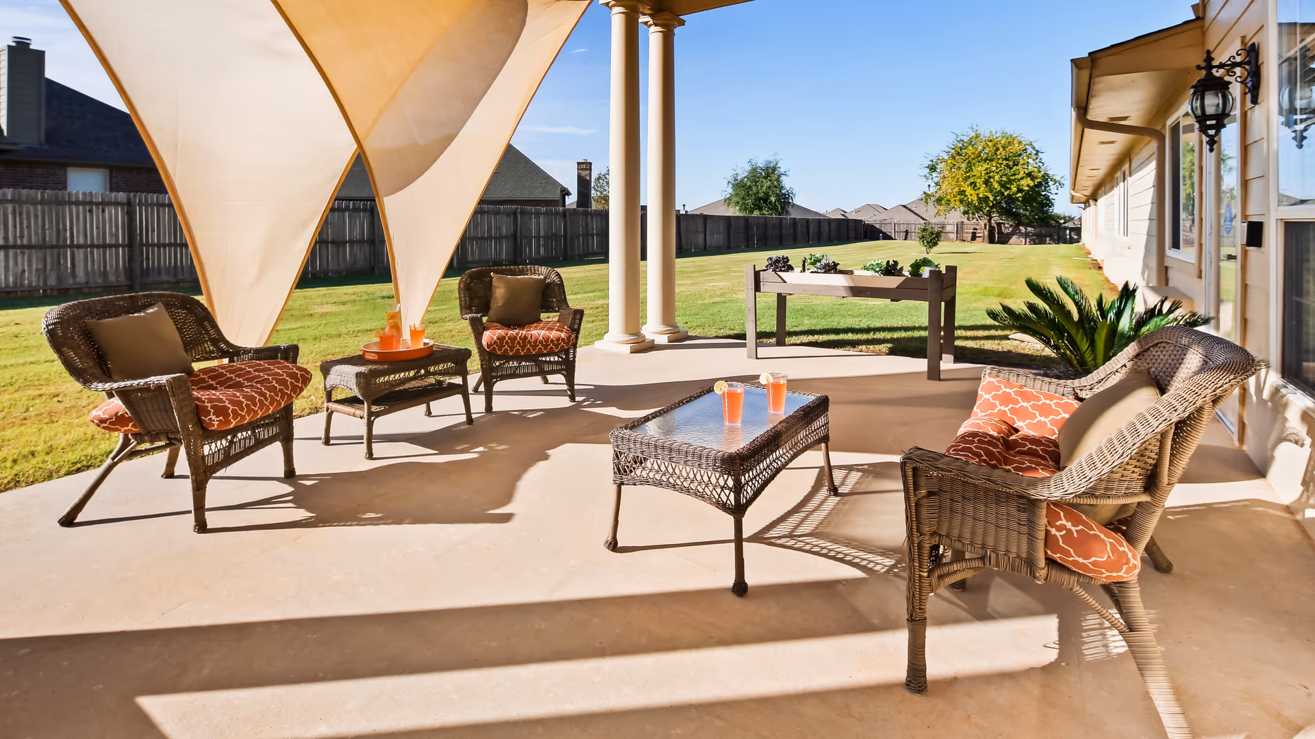 Outdoor patio area with wicker chairs and a wicker coffee table, each chair has patterned cushions. There are two triangular sun shades overhead, a small raised garden bed in the background, and a grassy yard enclosed by a wooden fence under a clear blue sky.