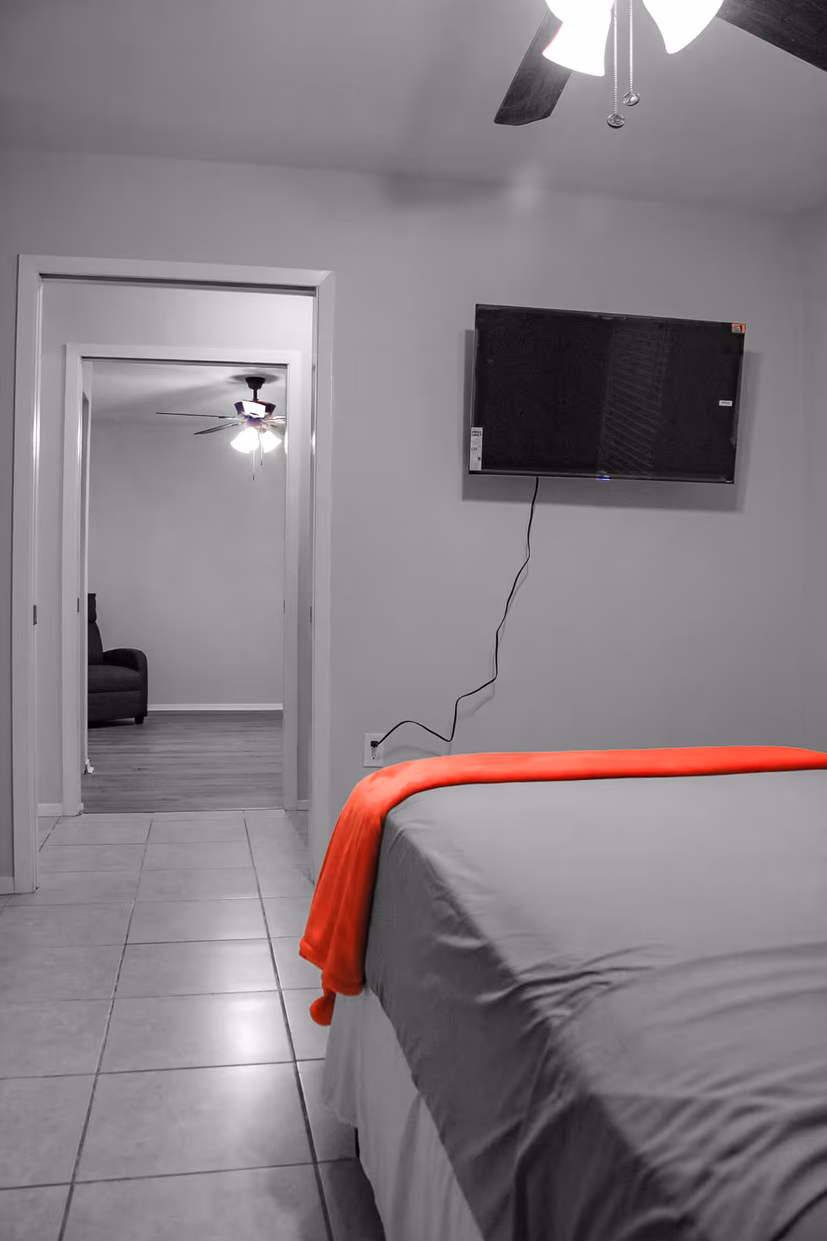 A bedroom with a bed and red throw, wall TV, doorway, and ceiling fan.