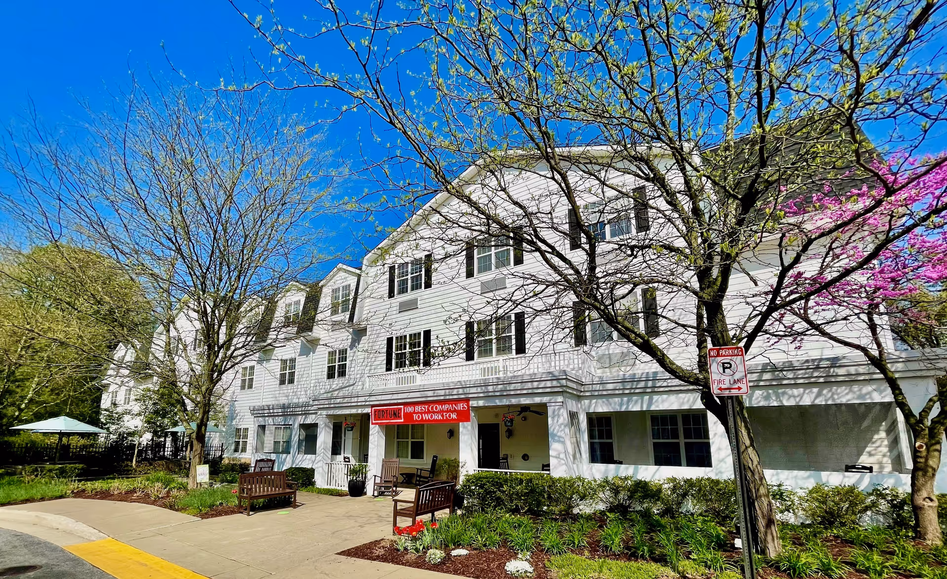 Exterior view of a three-story white senior living facility building with black window shutters, surrounded by trees with budding leaves and flowers under a clear blue sky. There are benches and a sidewalk in front of the building, along with a 'No Parking Fire Lane' sign.