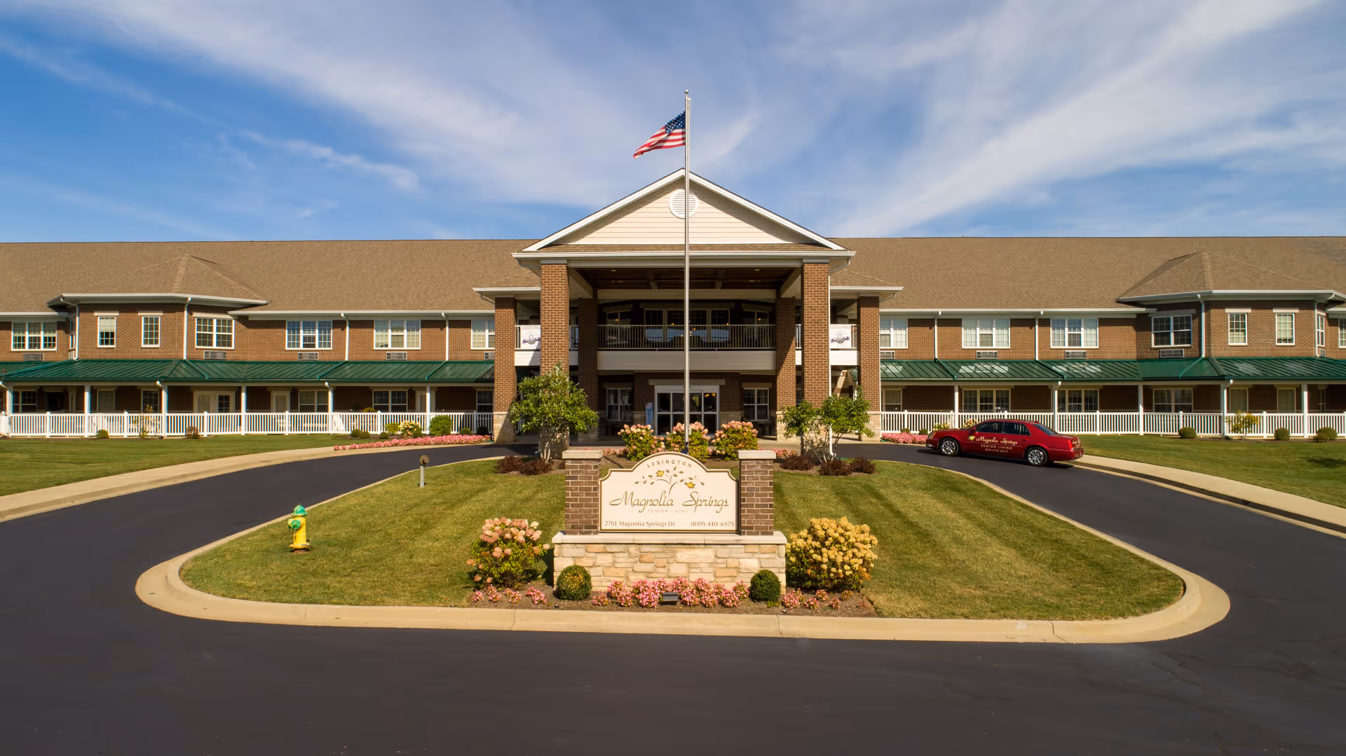 Front exterior view of Magnolia Springs Lexington, a large two-story brick building with a green roof over the porch area, an American flag on a flagpole in front, a landscaped lawn with flowers, and a driveway with a red car parked on the right side.