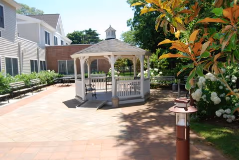 A white gazebo sits in a sunny brick courtyard with benches and lush greenery.